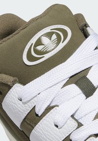 Olive green sneaker with white leather stripes, a textured upper, white laces, and an embossed logo on a circular patch.
