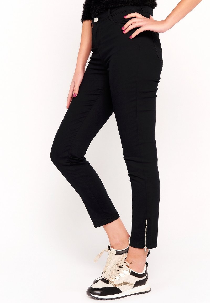 Mujer vestida con pantalones ajustados negros hasta el tobillo con cremalleras en los tobillos y zapatillas blancas y negras, posando con una mano en la cadera.