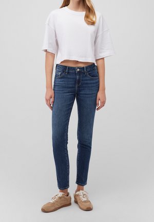 Kobieta w krótkim białym t-shircie, dopasowanych niebieskich dżinsach i beżowych sneakersach stoi na szarym, jednolitym tle.