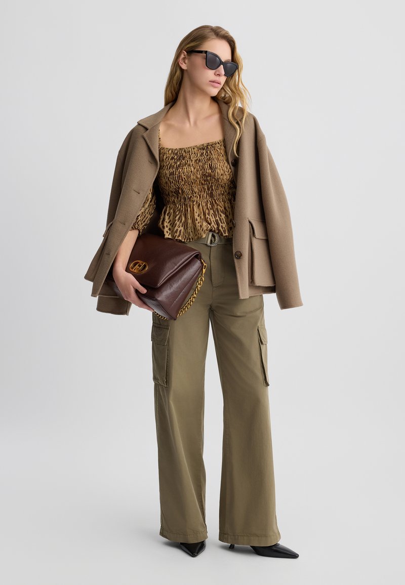 Top mit Leopardenmuster und Smock-Details, brauner oversized Mantel, olivgrüne Cargo-Hosen und eine braune Lederhandtasche mit goldener Kette. Schwarze Sonnenbrillen runden den Look ab.
