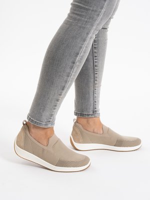 Füße in beigefarbenen, gestrickten Slip-on-Sneakern mit weißen Sohlen und grauen Skinny-Jeans vor weißem Hintergrund.