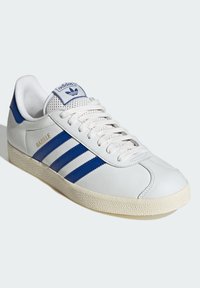 Bijele kožne tenisice s plavim trakama, strukturiranim gumeni potplat i podstavljenim jezikom. Sadrže logo i natpis "Gazelle" u zlatnoj boji sa strane.