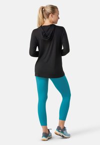 Svart långärmad topp med huva, parat med turkosa leggings och gråblå sportskor. Släta texturer och figurnära design.