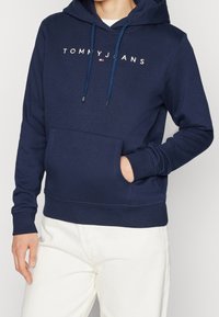 Sudadera de algodón azul marino con un bolsillo frontal, cordones en la capucha y el logotipo bordado en blanco "TOMMY JEANS" en el pecho.