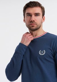 Camicia a maniche lunghe blu navy con scollo rotondo, caratterizzata da un bordino a righe rosse e bianche e un logo ricamato bianco sul petto. Tessuto morbido.