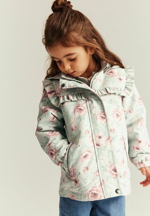 Junges Mädchen schaut nach unten, trägt eine hellblaue Blumenjacke mit Rüschen und blaue Jeans vor einem einfarbigen Hintergrund.