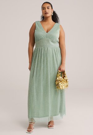 Kvinde iført en ærmeløs lysegrøn glitrende maxikjole, der holder en guldfarvet paliettasken og bærer hvide sandaler med stropper.