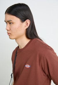Bruine katoenen t-shirt met ronde hals, voorzien van een klein, rond logo op de borst in rood, blauw en geel. Gladde textuur.