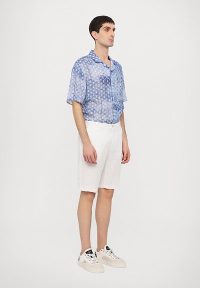 Chemise bleue à motifs à manches courtes associée à un short en lin blanc et des baskets blanches. La chemise présente une coupe décontractée et une fermeture à boutons.