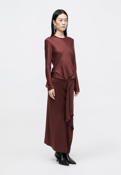 Vestido bordeaux de mangas longas com um detalhe frontal torcido, textura suave e drapeado lateral, combinado com botas de cano curto pretas com bico fino.