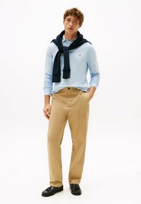 Jasnoniebieska koszula polo z długim rękawem, z granatowym swetrem zarzuconym na ramionach, w połączeniu z beżowymi chinosami i czarnymi mokasynami, wszystko na białym tle.