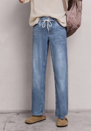 Person trägt hellblaue Jeans mit weitem Bein und weißem Kordelzug, beigen Strickpullover, beige Slip-On-Schuhe und hält einen mauvefarbenen Rucksack.