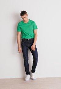 Zielona bawełniana koszulka z okrągłym dekoltem, ciemnoniebieskie dżinsy i białe trampki. Koszulka ma małe logo na piersi. Prosty, dopasowany krój.