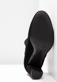 Zapato de tacón alto de ante negro con punta afilada y suela de goma texturizada. El tacón es cónico, ofreciendo una silueta elegante.