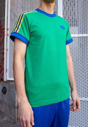 Grünes sportliches T-Shirt mit kurzen Ärmeln, blauem Kragen und Bündchen, mit gelbem Drei-Streifen-Design auf den Schultern und blauem Logo auf der Brust.