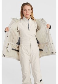 White zip-up jacket opened, revealing a light-colored high-neck top and matching pants. Textured fabrics and elastic waistband detail visible.
