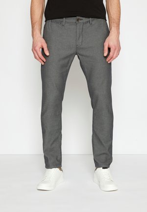 Mann trägt schmal geschnittene graue strukturierte Hose und weiße Sneakers, steht vor einem schlichten weißen Hintergrund.