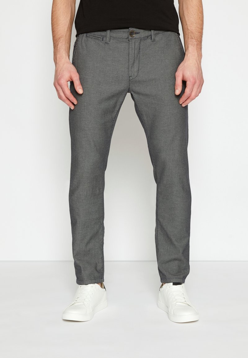 Homme portant un pantalon gris ajusté texturé et des baskets blanches, debout devant un fond blanc uni.