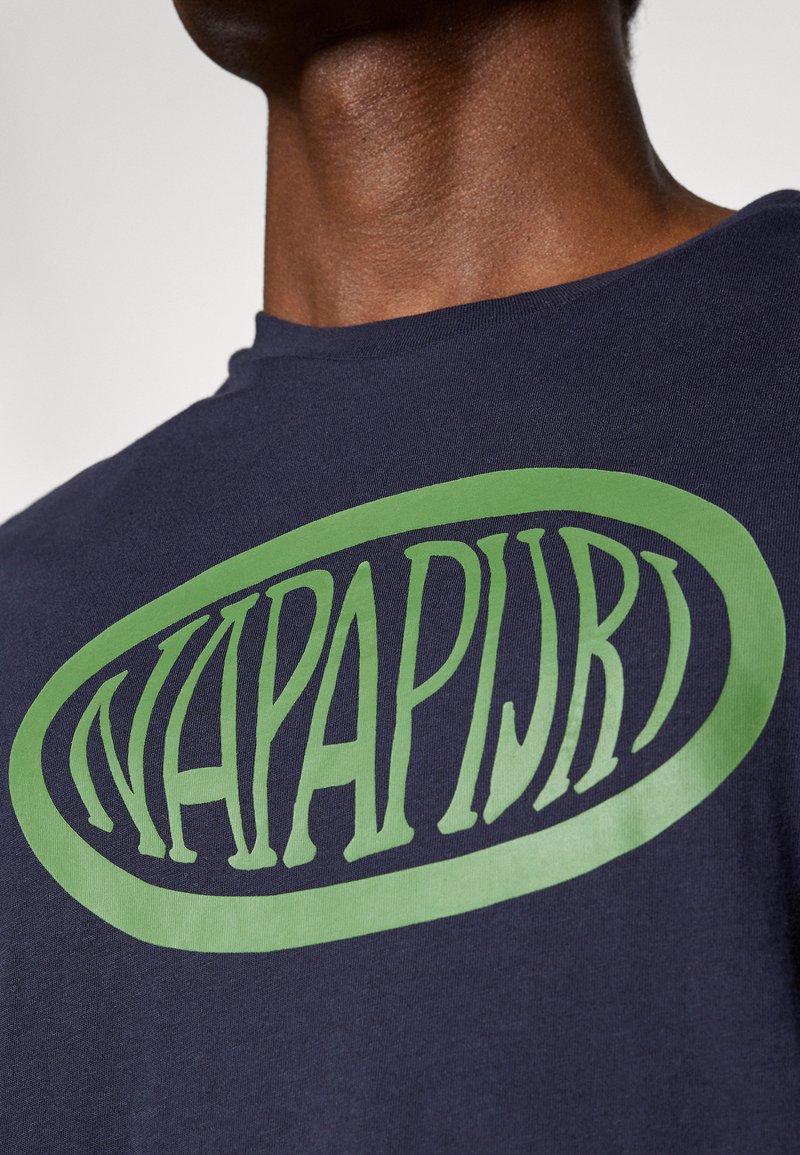 T-shirt bleu marine avec un grand logo ovale vert portant le texte "NAPAPIJRI" dans une police stylisée, fabriqué en tissu coton.