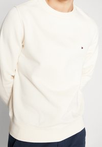 Sudadera de cuello redondo color crema, elaborada con un tejido suave; presenta un pequeño logo en rojo, blanco y azul en el lado izquierdo del pecho; puños y dobladillo acanalados.