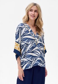 Bluzka z krótkim rękawem z białego materiału z niebieskim wzorem liści i złotymi akcentami wzdłuż dekoltu i rękawów. Projekt z dekoltem w kształcie litery V.