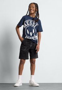 Marineblå t-skjorte med hvit "Messi"-logo, kombinert med sorte knelang shorts og hvite joggesko. Flettet frisyre og selvsikker holdning.