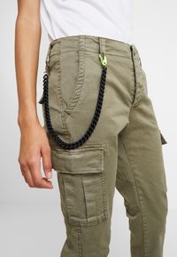 Olijfgroene cargobroek met zijzakken, geweven stof, zwarte kettingdetails bevestigd met een neon groene haak op de tailleband.