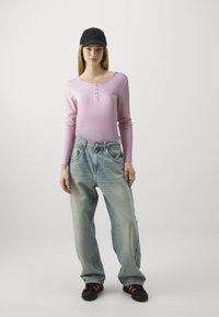 Top lungo a maniche ribattute rosa con dettagli di bottoni, abbinato a jeans larghi azzurri chiari. Un cappello nero e sneakers nere con accenti rossi completano l'outfit.