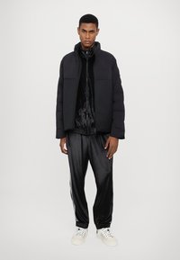 Black puffer jacket with high collar, zip front, and side pockets, layered over a shiny black track jacket and matching track pants, worn with white sneakers.