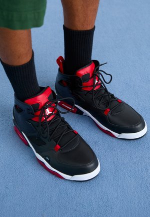 Czarne, czerwone i białe wysokie sneakersy noszone z czarnymi skarpetami na jasnoniebieskiej podłodze, pokazujące stopy i dolne części nóg w zielonych szortach.