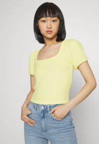 Top amarelo com franzidos, de mangas curtas e decote quadrado, com um design justo e ligeiras mangas bufantes, combinado com uns jeans azuis claros.