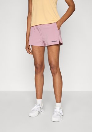 Rosa Sportshorts mit Seitentaschen und einem Logo am linken Bein, kombiniert mit einem hellgelben Oberteil und weißen Turnschuhen.