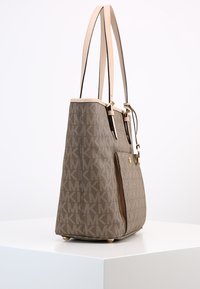 Sac fourre-tout marron avec motif de logo, accents en cuir beige, forme structurée, double anses et quincaillerie en métal. Surface texturée et base plate.