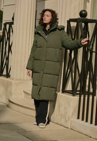 Geacă puffer verde olive cu glugă, având închidere cu nasturi și design matlasat. Purtată cu pantaloni bleumarin și teniși albi, în aer liber, pe scări.
