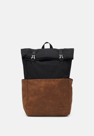 Sac à dos à rabat avec partie supérieure en tissu noir et partie inférieure en cuir marron. Équipé de quincaillerie métallique et d'une poignée supérieure pour le transport.