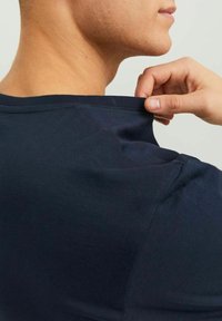 Tricou din bumbac de culoare albastru marin, cu decolteu rotund, având o textură netedă și o întindere subtilă, vizibilă la gât, unde este ușor tras.