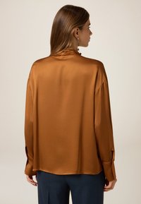 Brązowa satynowa bluzka z wysokim kołnierzem i luźnym krojem. Posiada długie rękawy z guzikami oraz boczne rozcięcia dla ułatwienia ruchów.