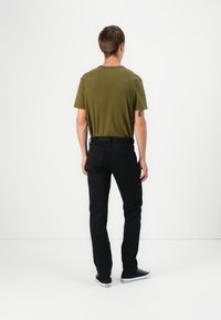 Hombre de pie de espaldas, vistiendo una camiseta verde oliva, jeans negros y zapatillas Converse negras, frente a un fondo blanco liso.