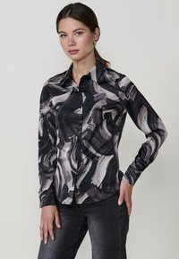 Blusa a maniche lunghe nera con motivi astratti a pennellate grigie. Presenta un colletto classico e chiusura frontale con bottoni. Tessuto dalla texture liscia.