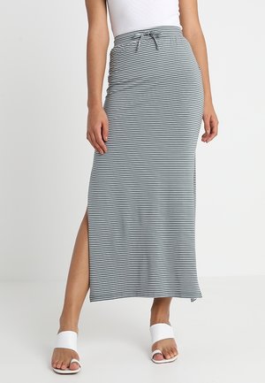 Femme portant une jupe maxi rayée horizontale grise et blanche avec une fente sur le côté, un haut blanc et des sandales à bout ouvert blanches.