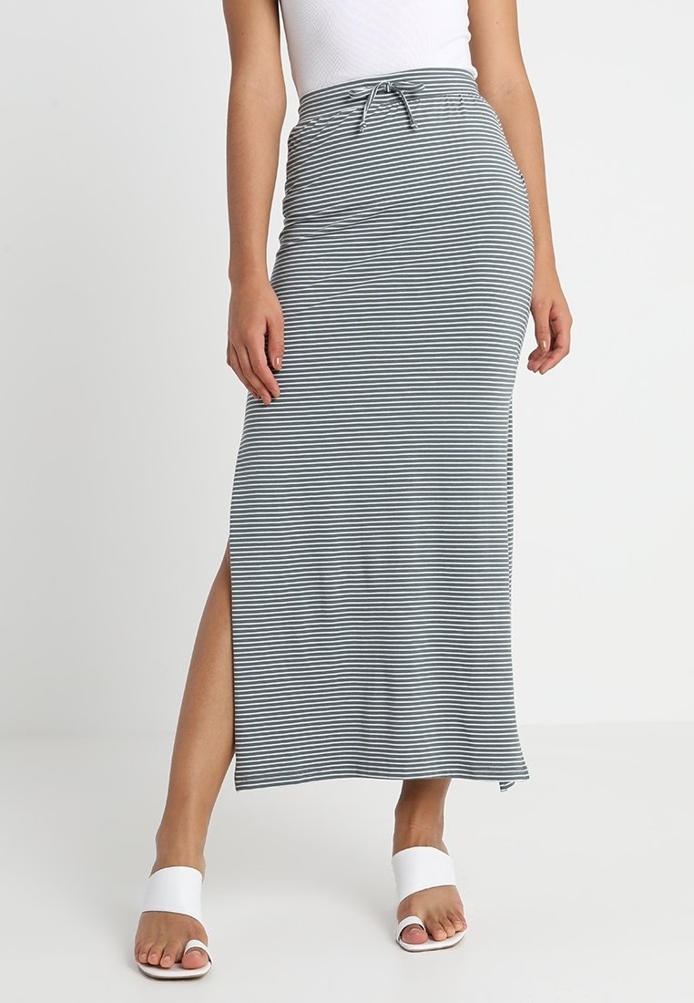 Femme portant une jupe maxi rayée horizontale grise et blanche avec une fente sur le côté, un haut blanc et des sandales à bout ouvert blanches.