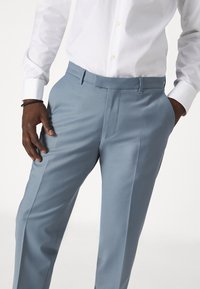 Pantalon bleu clair avec une texture lisse, coupe ajustée, devant plat et poches latérales, associé à une chemise blanche à boutons.