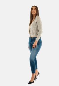 Cardigan argento scintillante con scollo a V e bottoni, abbinato a jeans skinny blu scuro e tacchi neri. Texture lisce e design aderente.