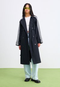 Manteau noir en trench avec un design à double boutonnage, des manches rayées et une coupe décontractée, porté sur un top blanc court et un jean bleu clair.