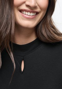 Sort top med høj hals og nøglehul-detail, lavet af et glat stof. Model med langt brunt hår og en neutral baggrund.