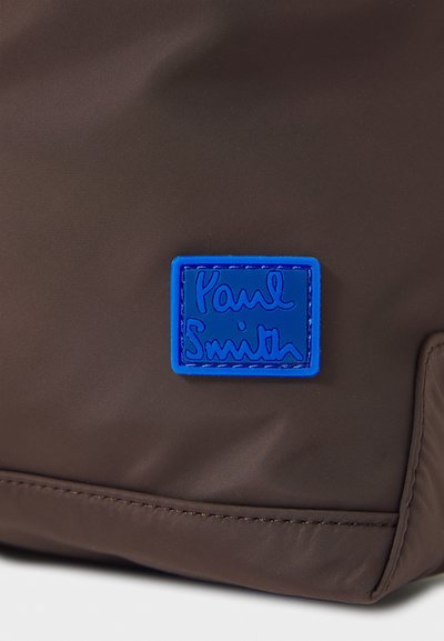 Pruun nailonist kott, millel on sinine tekstuuriga silt "Paul Smith" logoga, ristkülikukujuline, sile pind ja õmblemise detailid.