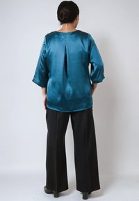 Woman with dark hair in a bun wearing a shiny teal 3/4 sleeve blouse and black wide-leg pants, standing against a plain light background.