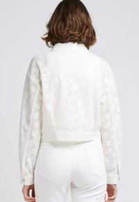 Veste blanche courte en tissu semi-transparent à motifs floraux avec des poignets boutonnés. Associée à un pantalon en denim blanc avec des poches arrière.