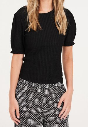 Femme aux cheveux blonds ondulés portant un haut noir texturé à manches courtes et un pantalon noir et blanc à motifs, debout, les mains détendues le long du corps.