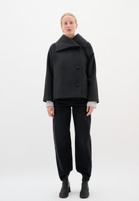 Schwarze Wolljacke mit weitem Kragen und zwei großen vorderen Knöpfen, getragen über einem hellgrauen langärmeligen Hemd und kombiniert mit schwarzen weiter geschnittenen Hosen.